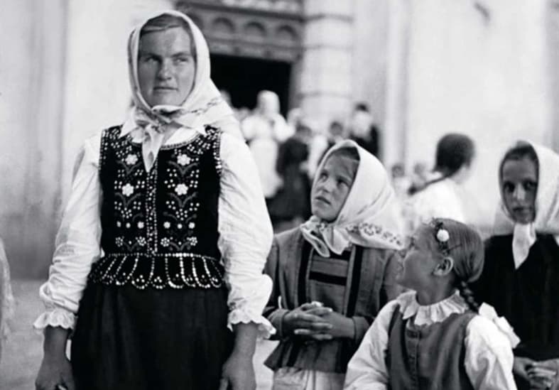 Дівчина у святковому одязі стоїть під церквою у Сокільниках (село Пустомитівського району Львівської обл.), чекаючи на приятеля. Молодша дівчинка прикрасила волосся живими квітами